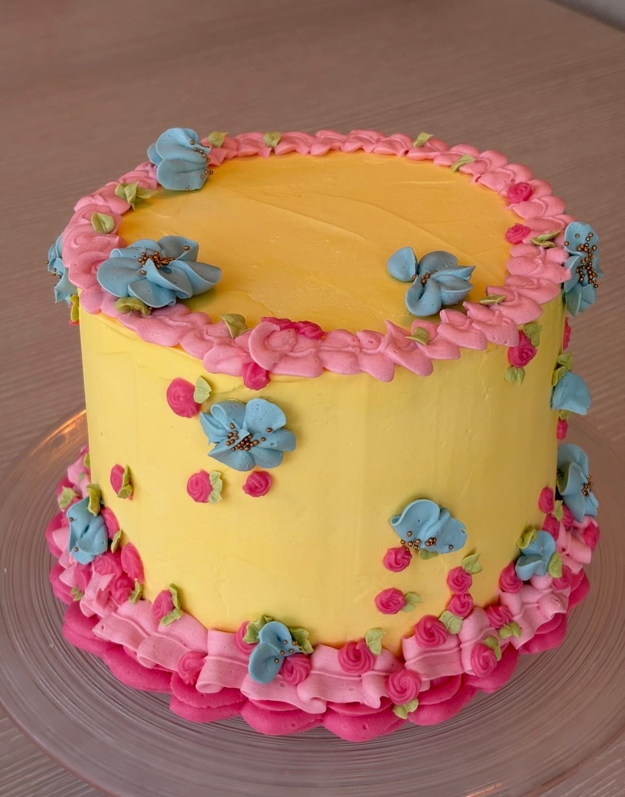 Gul lagdelt smørkremkake med rosa og blå blomster og roserosetter rundt toppen og bunnen